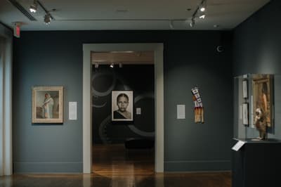 Art gallery with portraits, a fabric piece, and a sculpture displayed against dark gray walls.