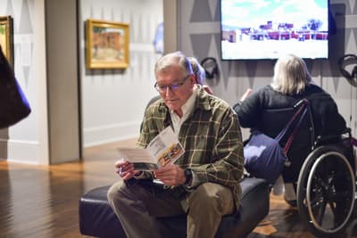Seated gentleman o bech in art gallery reading event program.