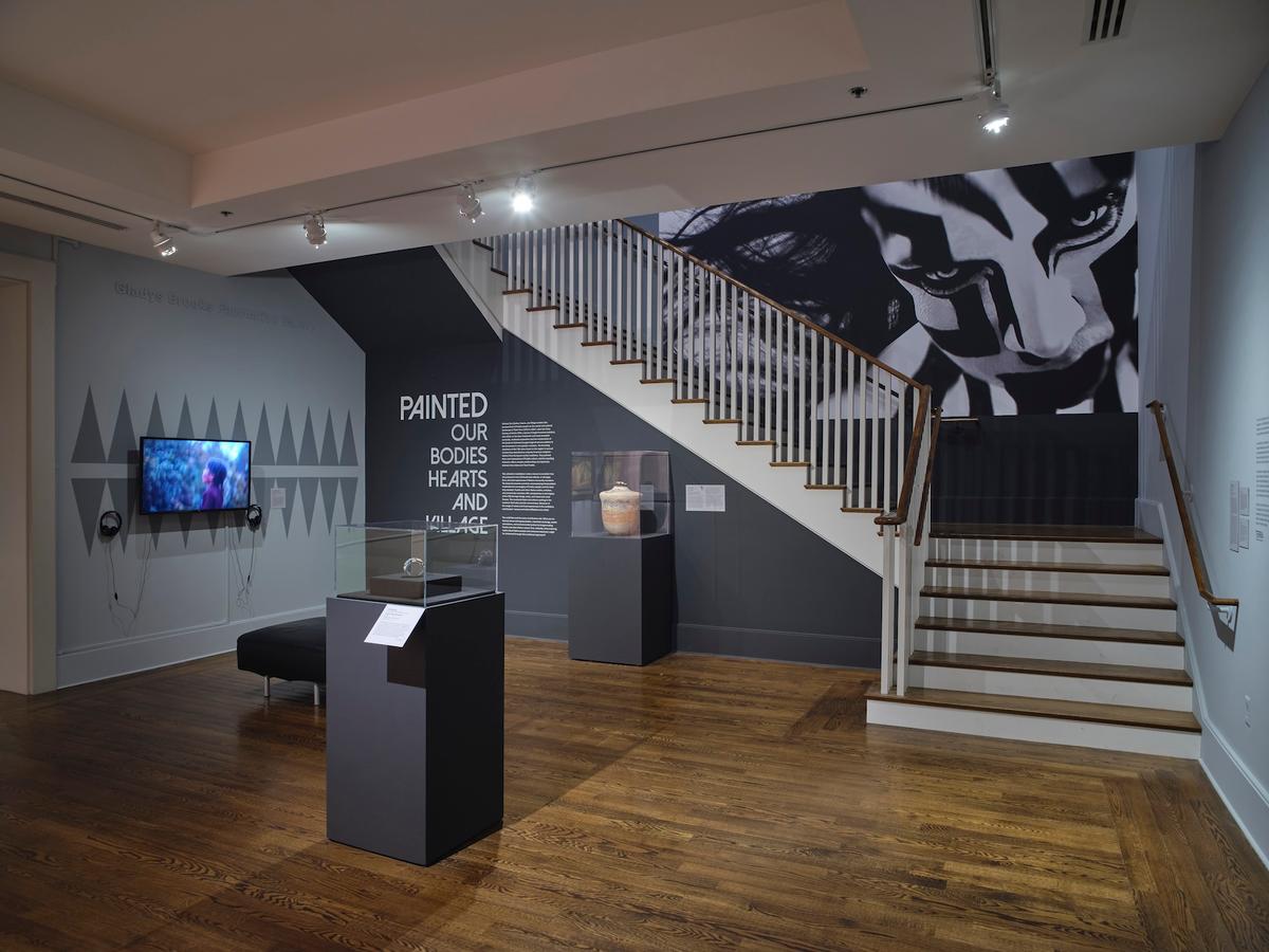 Gallery space with exhibition title wall reading "Painted: Our Bodies, Hearts, and Village" beside a staircase. The room includes display cases on pedestals and a large black and white photograph protrait hung in the staircase.