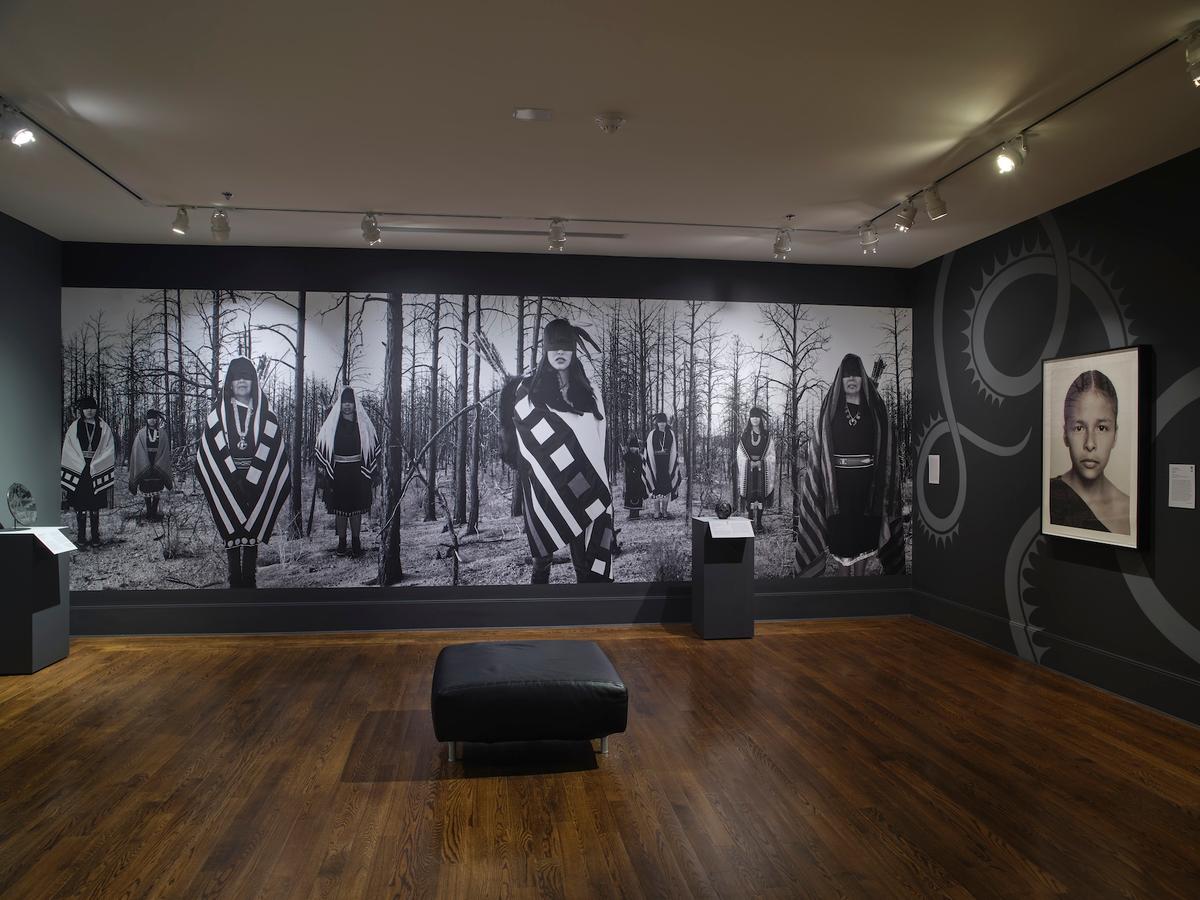 A gallery with a large black and white photograph of nine figures standing blindfolded in the woods, alongside a portrait on the adjacent wall.