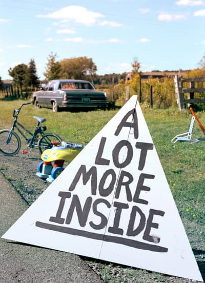 A three-dimensional triangular object outside along a driveway and grassy area with hand-painted lettering that reads A LOT MORE INSDE.