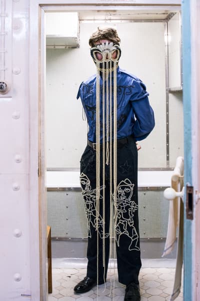 Person in a mask with long strings standing in a doorway, wearing a blue shirt and patterned pants.