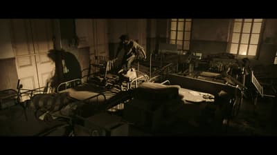 A film still in which a figure in motion moves through an abandoned room with bedframes and rusty furniture.