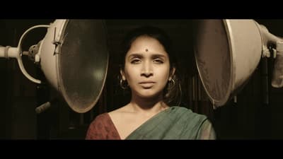 Still from a film. A woman wearing a sari and bindi is central to the shot, and two large medical lights are on either side of her head.