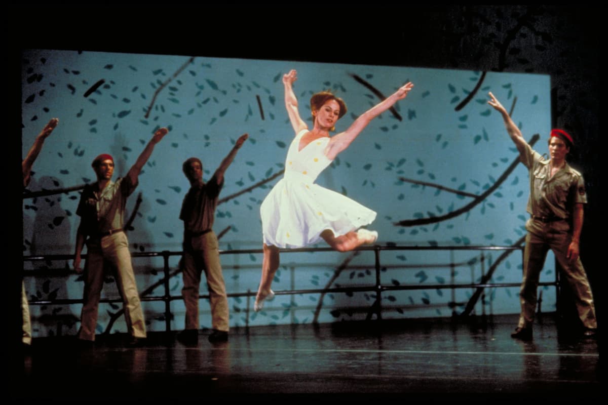 A female dancer in a white dress leaps on stage, surrounded by male dancers in khaki uniforms with a leafy backdrop.