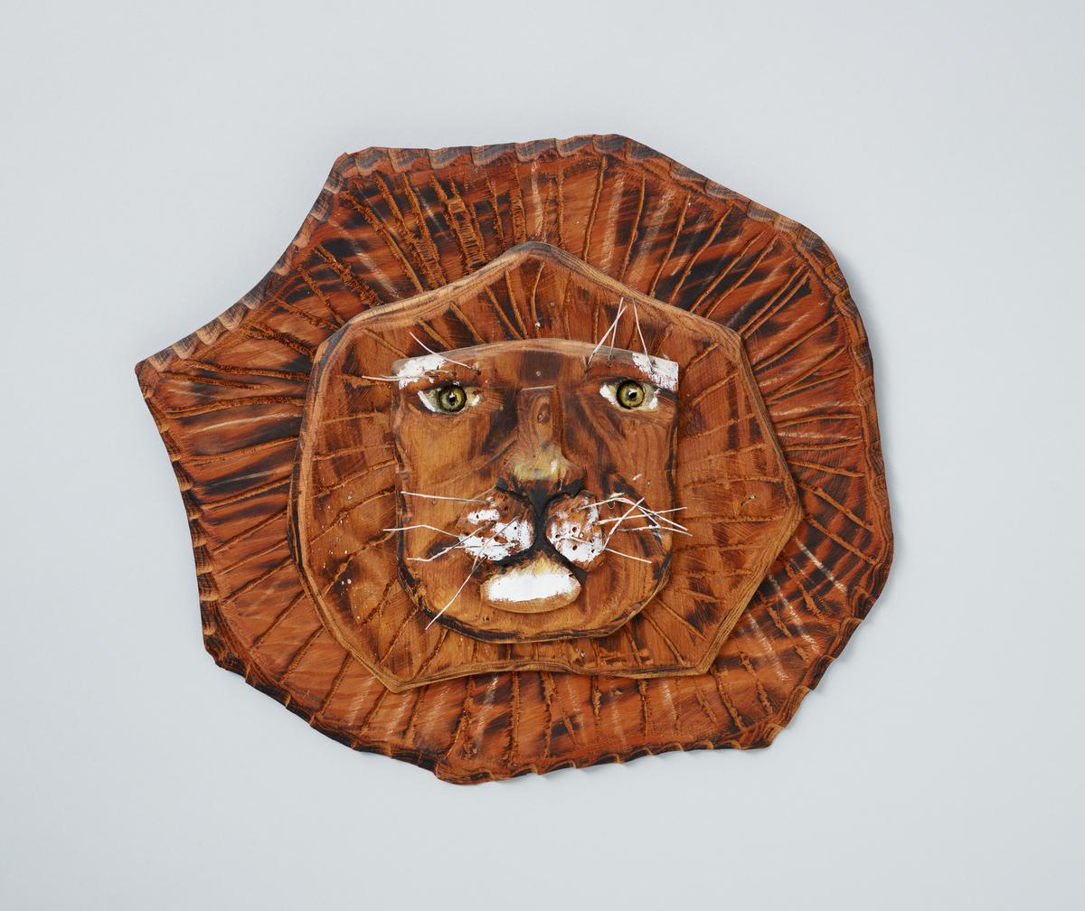 A carved, stained, painted, and blackened wood sculpture of a lion's face and mane with wire whiskers.