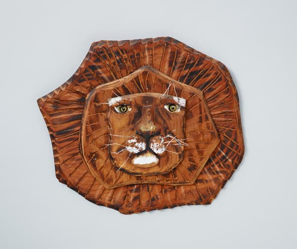 A carved, stained, painted, and blackened wood sculpture of a lion's face and mane with wire whiskers.