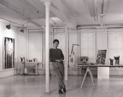 A black and white photograph of the artist Martha Diamond leaning against a column in her large, well-lit studio. She has short hair, her hands are folded in front of her, and she is meeting the gaze of the camera intently. Paintings are on the walls and brushes and other tools are on a cart and on the table. The space is clean, spare, and organized.
