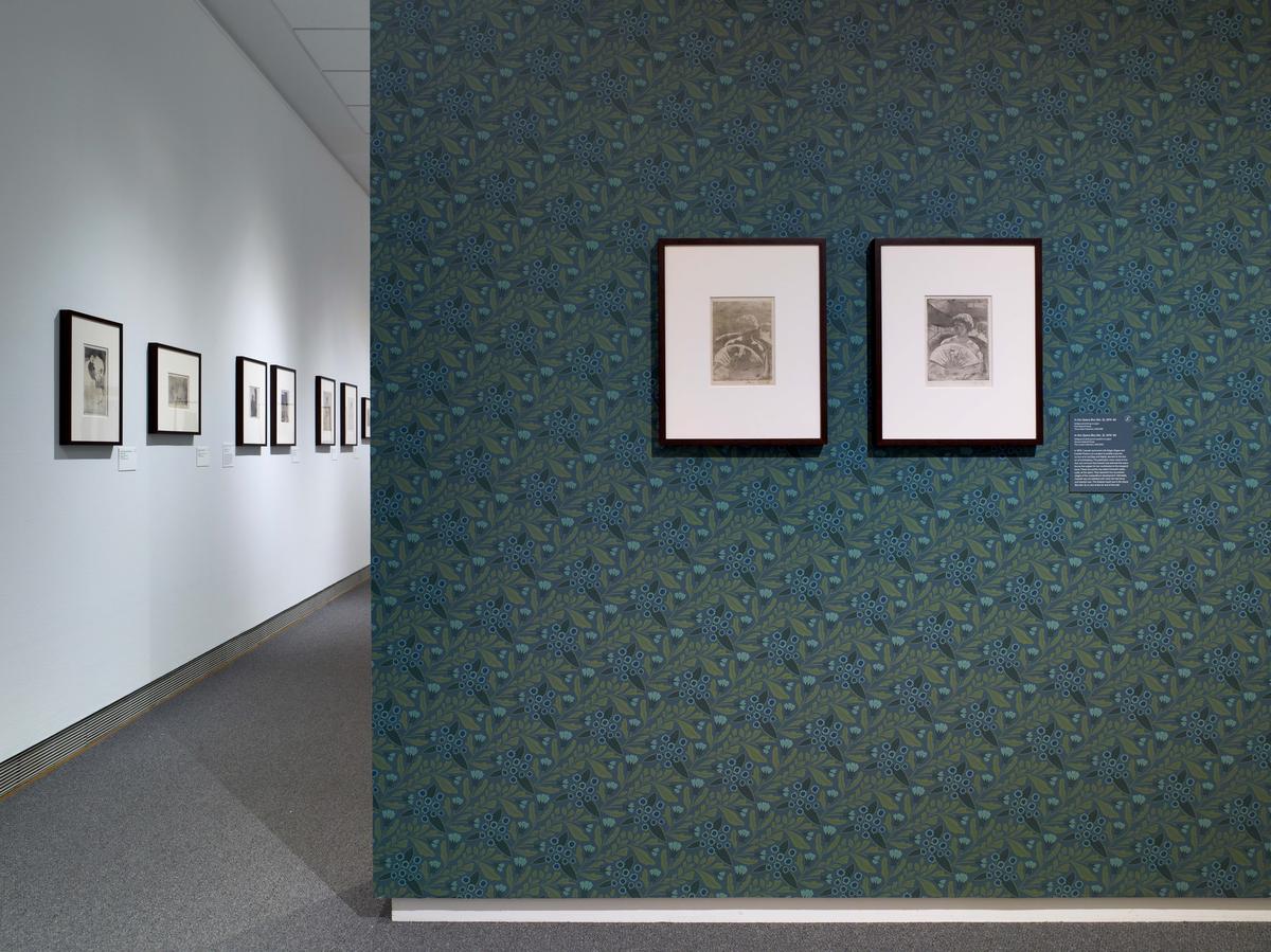A gallery space with framed artworks on a white wall and a patterned wall with floral designs.