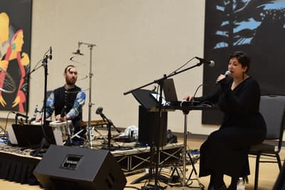 Seated woman sings into microphone while someone plays drums