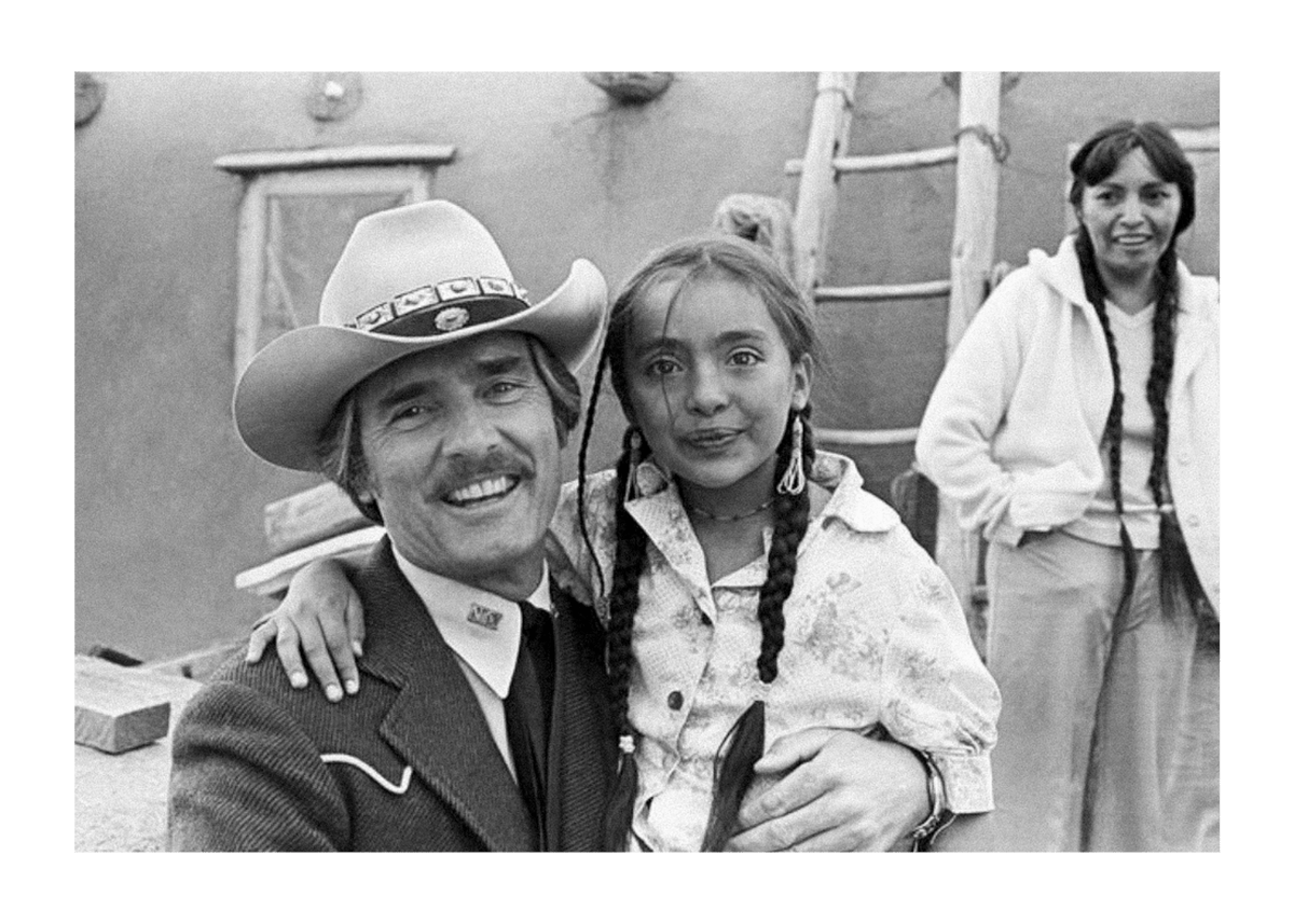 A man in a cowboy hat holds a smiling young girl with braided hair, while a woman stands in the background next to a rustic building with a ladder.