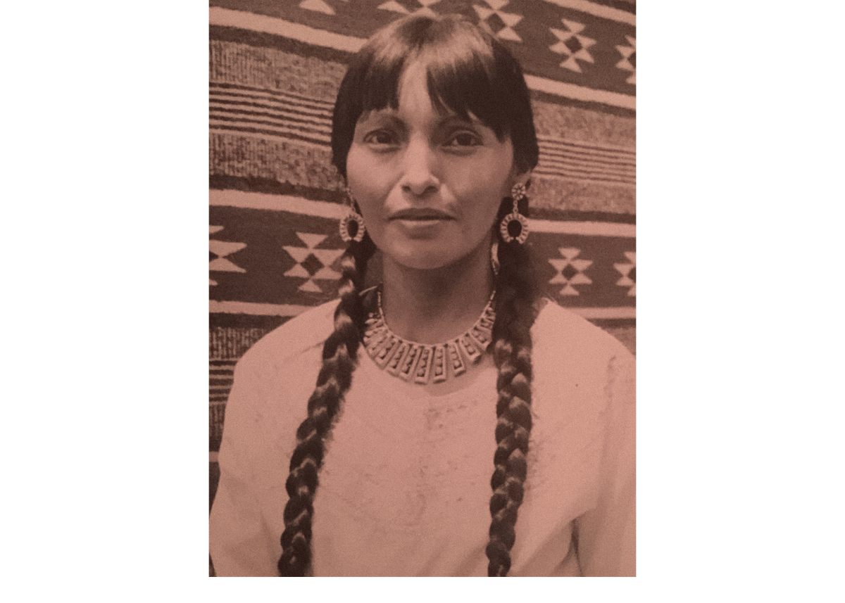 Sepia-toned portrait of a woman with long braids, wearing a necklace and earrings, against a patterned textile background.