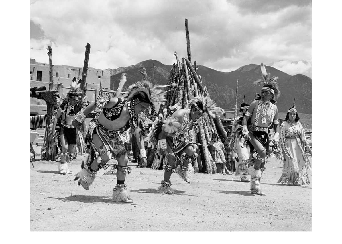 Native American dancers in traditional regalia performing in front of mountains.