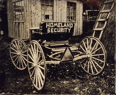 An old wooden cart with "HOMELAND SECURITY" painted on the back, in front of a rustic building.