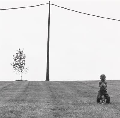 Photograph, a little boy sits atop a mannequin head in empty grassy field, with tree in far left background and wired utility pole overhead.