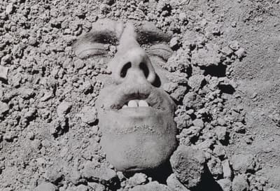 A black and white photographic self-portrait by David Wojnarowicz with his face buried in the dirt, 1991/2018.