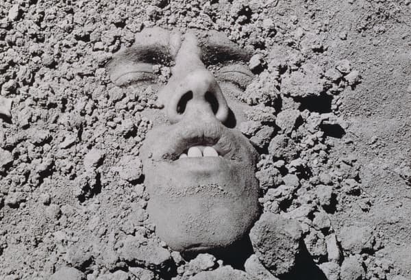 A black and white photographic self-portrait by David Wojnarowicz with his face buried in the dirt, 1991/2018.