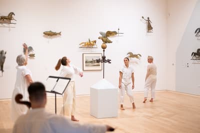 Four dancers in motion in front of a wall of art accompanied by a cellist