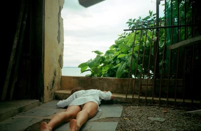 Person lying face down on a tiled floor in a partially open structure with plants and a view of the water in the background.