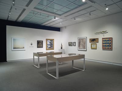 An art gallery with white walls displaying various artworks and two glass display tables.