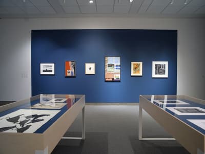 An art gallery room with a blue accent wall displaying framed artworks and two display tables with prints.