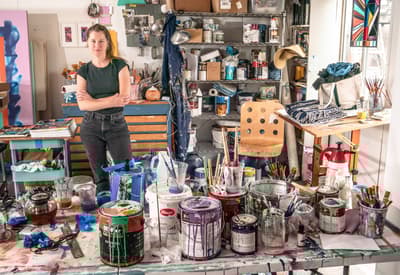 Artist standing in a cluttered studio with paint supplies and tools scattered on a table.