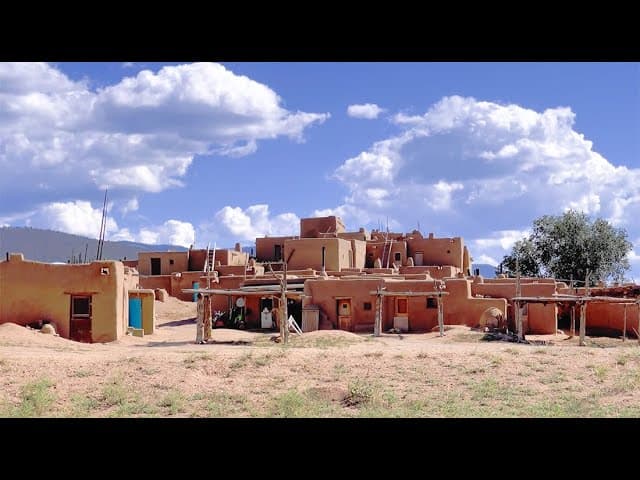 Traditional Pueblo village with adobe structures and wooden ladders under a blue sky with clouds.