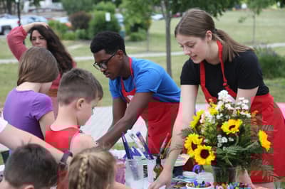 Adults and children engaging in an outdoor art activity with flowers and paint supplies on the table.