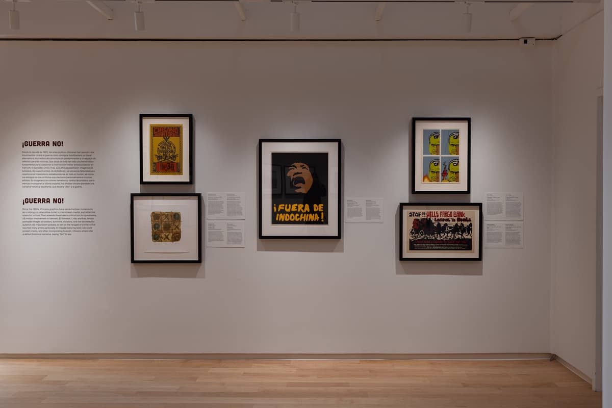 Wall display of Chicanx protest graphics with posters opposing war, featuring bold colors and text in Spanish and English under the heading “¡Guerra No!”