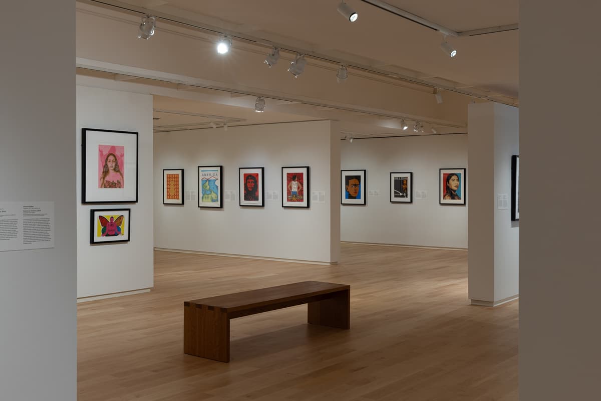 Wide view of the gallery space featuring colorful framed portraits, posters, and prints with a wooden bench placed centrally on the wooden floor