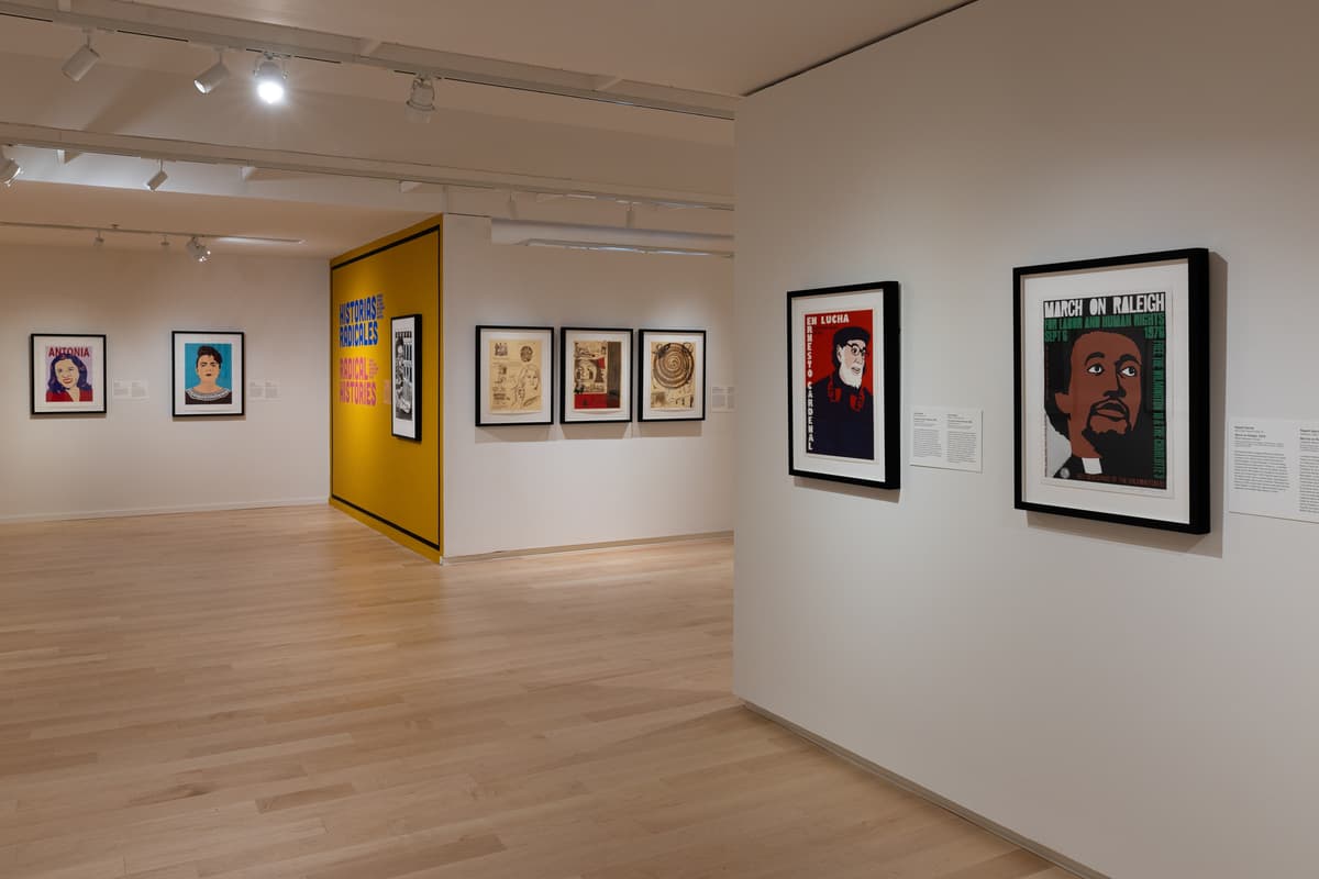 Gallery view with framed protest prints along three walls and the “Radical Histories” exhibition title in bright colors on a central yellow partition