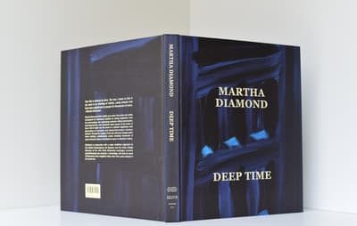 A photo taken from the angle of the book binding of a book propped up. The book cover has a full bleed abstract blue painting with the text MARTHA DIAMOND DEEP TIME in a light yellow
