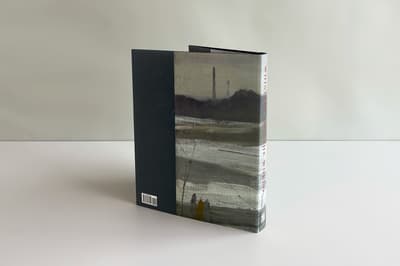 Back cover of a book propped up. Two thirds vertically of the back cover are of a gray painting depicting a river scene, and the other one third is navy