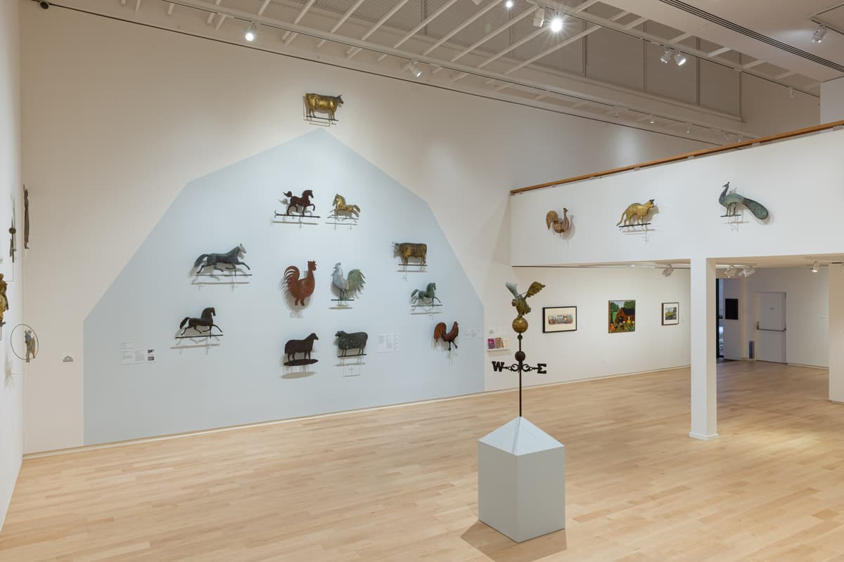 Animal-shaped weathervanes mounted on a wall with a pale blue outline mimicking a roofline. In the foreground, an eagle weathervane sculpture installed on a plinth.