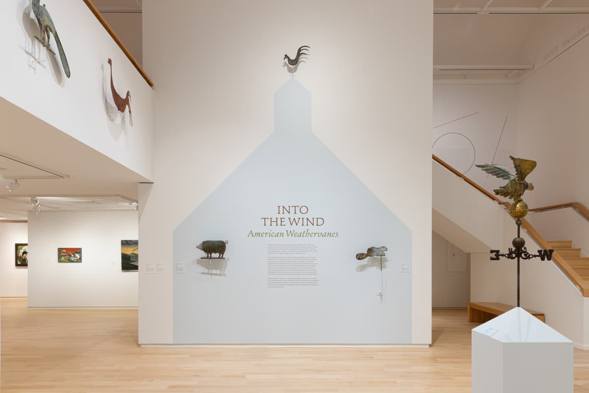 Wall text reading “Into the Wind: American Weathervanes” framed by mounted weathervanes shaped like animals, and a large eagle weathervane in the foreground on a plinth.