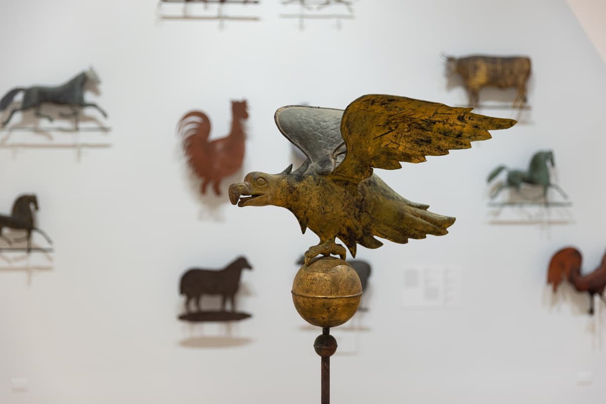 Close-up of a gilded eagle weathervane in the round, with a backdrop of various animal-shaped weathervanes hung on a wall.