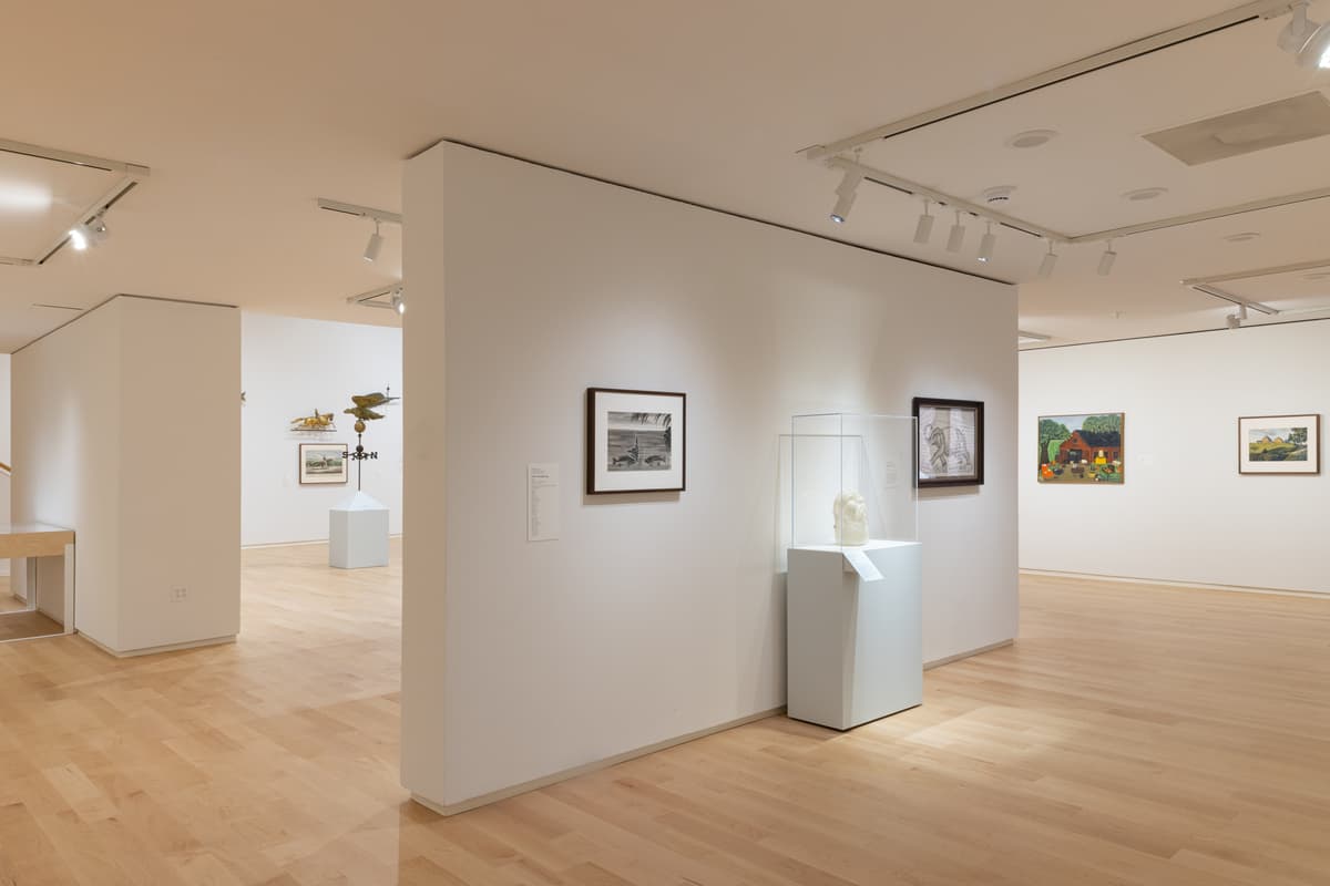 Gallery view with paintings on the walls, and a glass-encased sculpture on a pedestal in the center; weathervanes visible in the adjoining space.