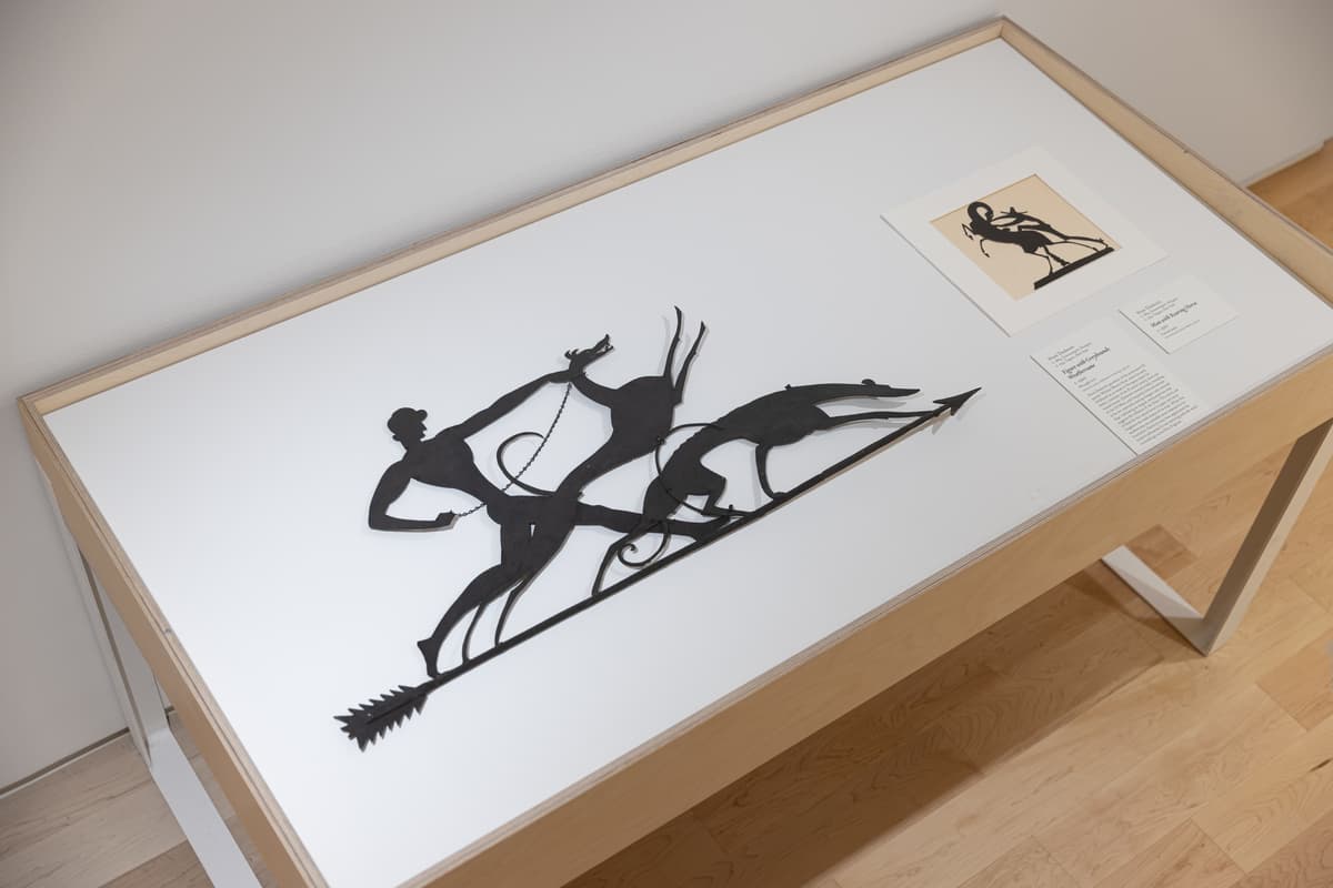 Vitrine with weathervane depicting a hunter with bow and arrow pursuing a deer, displayed in a vitrine with accompanying interpretive texts and archival image.