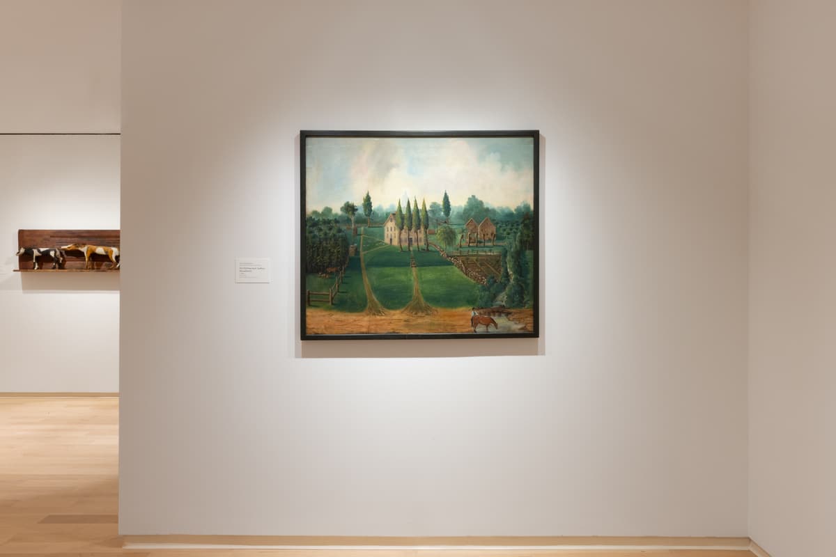 Framed folk-style landscape painting depicting a lush green countryside with buildings and animals, hung on a gallery wall with spot lighting.