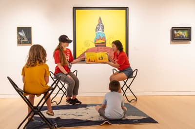 Two adults holding a book, reading to two children in an art gallery setting with paintings on the wall.