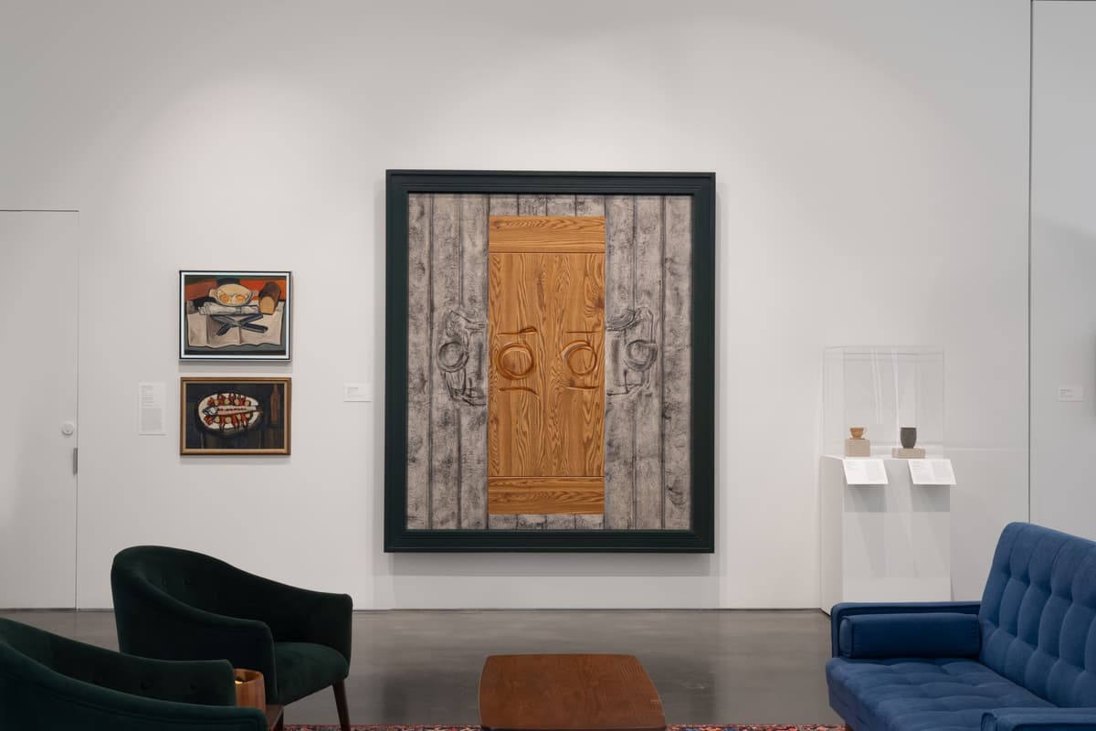 Large framed painting depicting a wooden table set with two plates from a bird's eye view with an abstraction of one individual on each side. Artwork is flanked by still-life paintings and a display case with pottery, with blue and green seating in the foreground