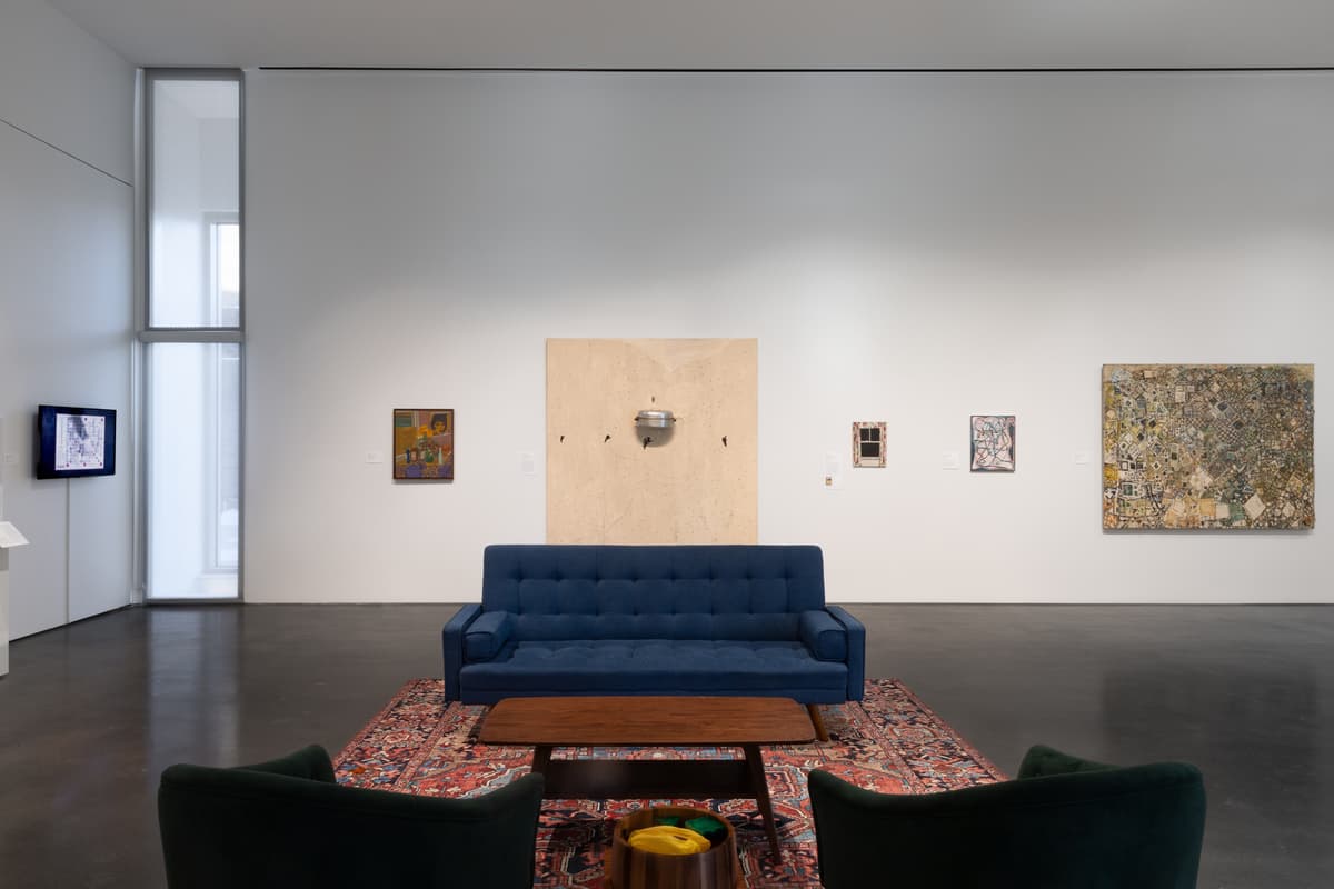 Seating area with blue and green furniture, centered in front of a large wall displaying a wooden panel with a small metal object, surrounded by various paintings
