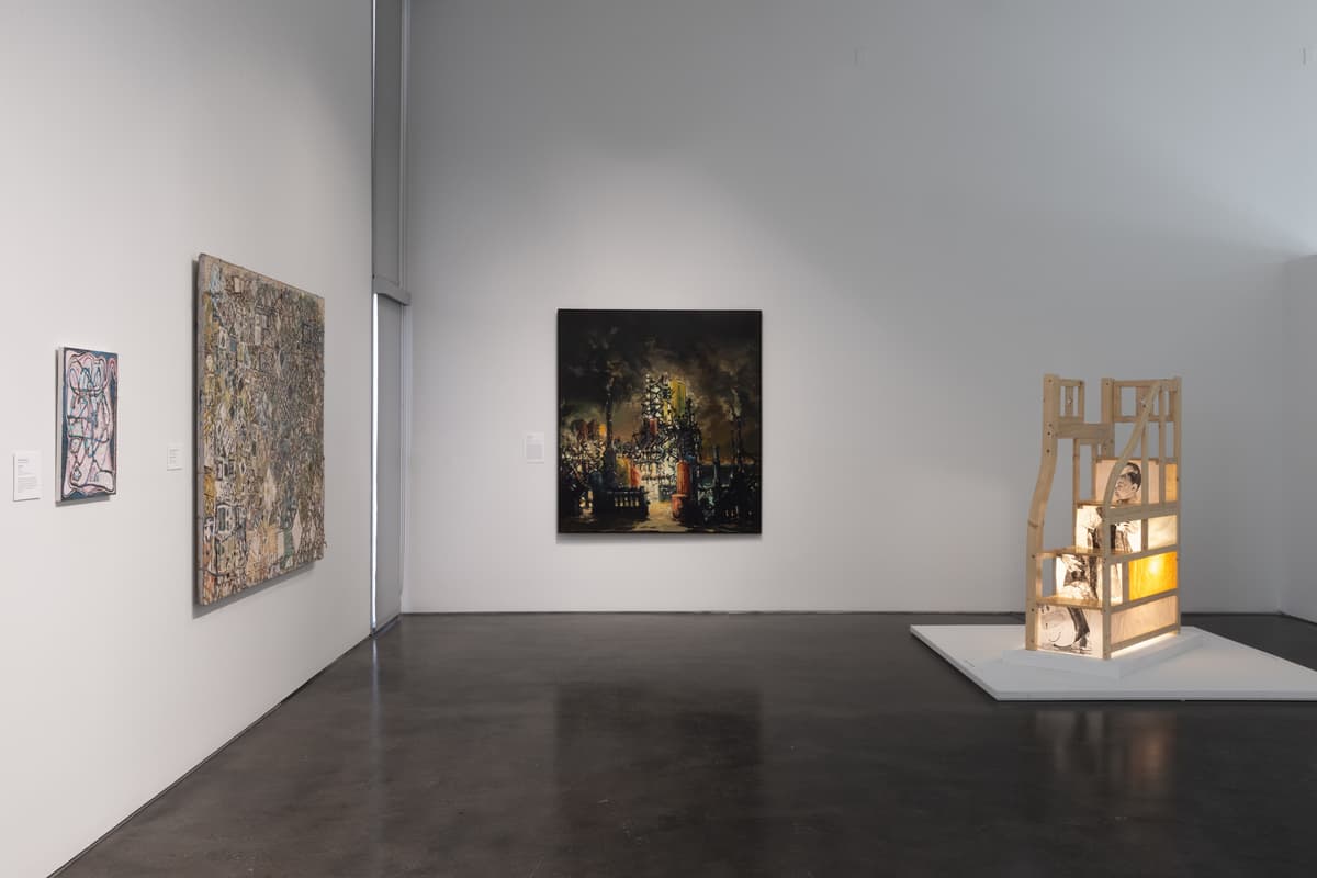 Corner view of the gallery showing an illuminated wooden sculpture on a platform, with a dark atmospheric painting on the wall and two abstract works on the left