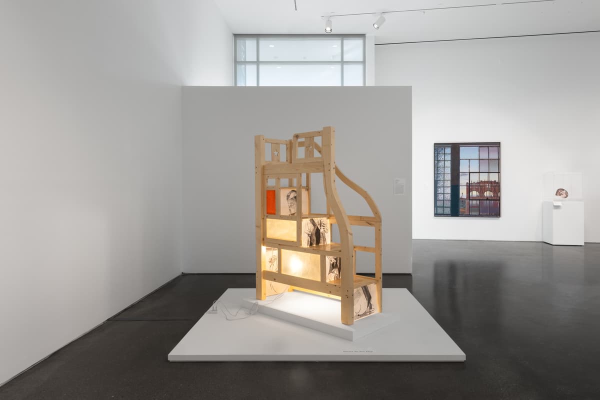 A glowing, wooden-framed sculpture with illustrated panels on a platform, shown from the rear, with additional artworks visible in the background