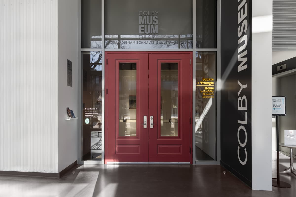 Entrance to the Colby Museum’s Joan Dignam Schmaltz Gallery of Art, with red double doors and signage for the exhibition “Square + Triangle: Home in the Colby Museum’s Collection”