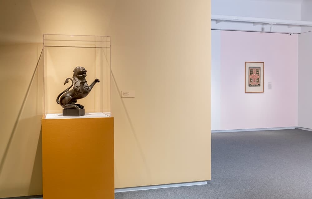 Bronze animal sculpture in a display case on a yellow pedestal, with framed artwork on a white wall in the background