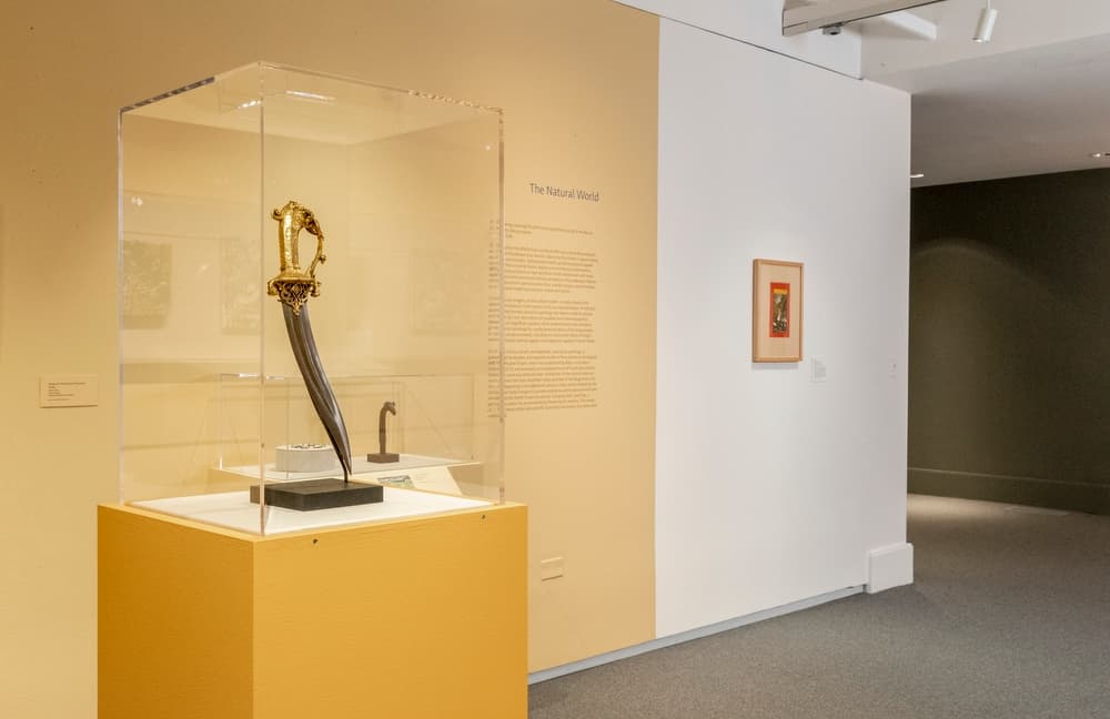 Ornate gold-handled sword displayed upright in a clear vitrine against a pale yellow wall with wall text