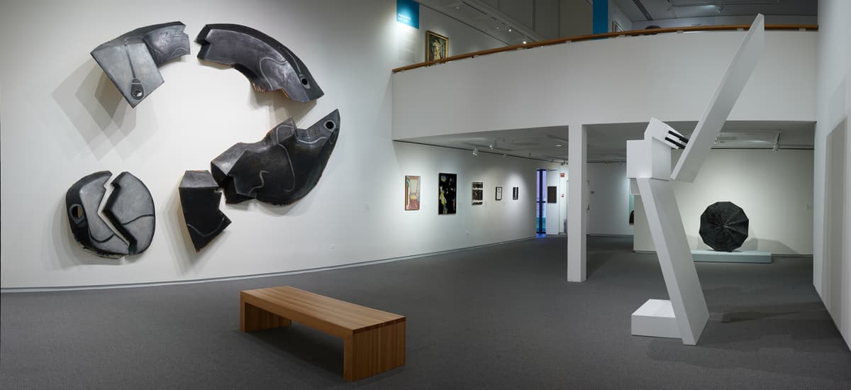 A panoramic view featuring a ceramic wall sculpture, freestanding geometric sculpture, and bench in foreground with several wall hung works visible in the background.