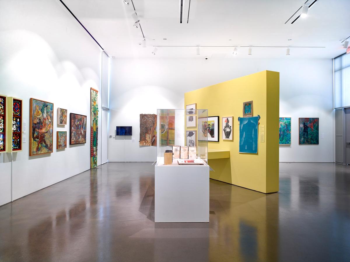 Gallery installation with colorful paintings on white walls, a yellow central partition, and a vitrine of books.