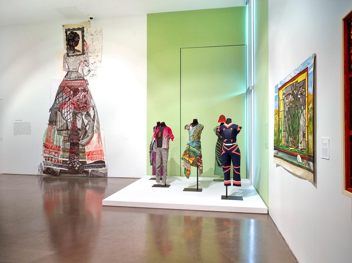 Five mannequins in vibrant garments on a platform surrounded by other works of art.